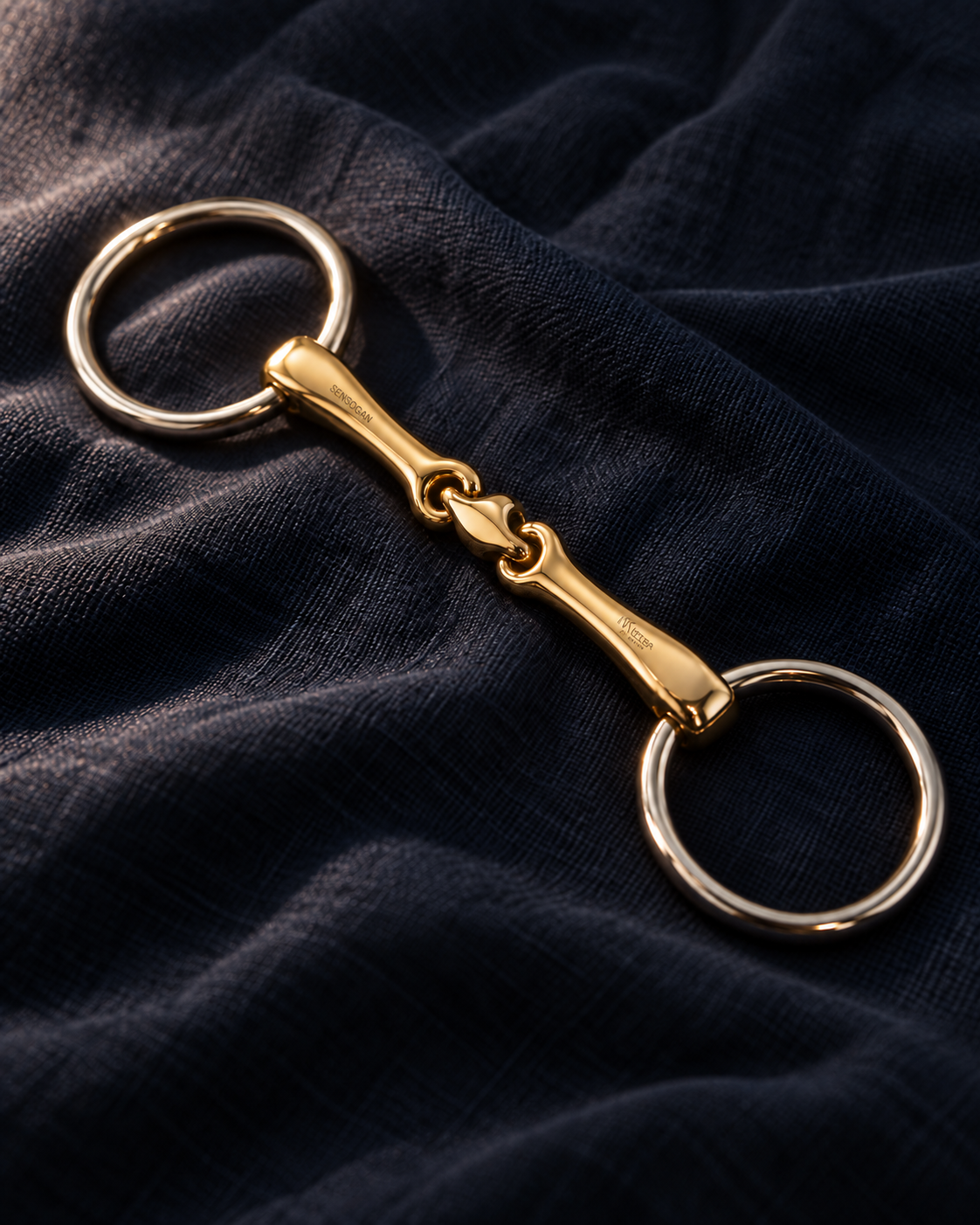 Loose ring snaffle bradoon on navy linen, polished silver rings and a warm gold double-jointed mouthpiece with a central link