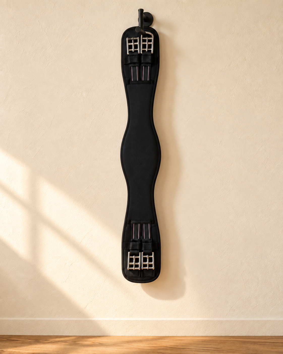 Black padded dressage girth hanging full length from an iron hook on a cream plaster wall, with two stainless roller buckles and black elastic at each end