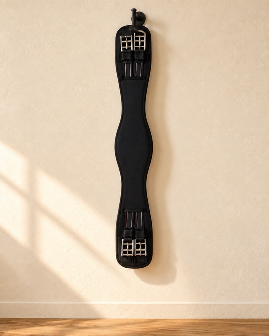 Black padded dressage girth hanging full length from an iron hook on a cream plaster wall, with two stainless roller buckles and black elastic at each end