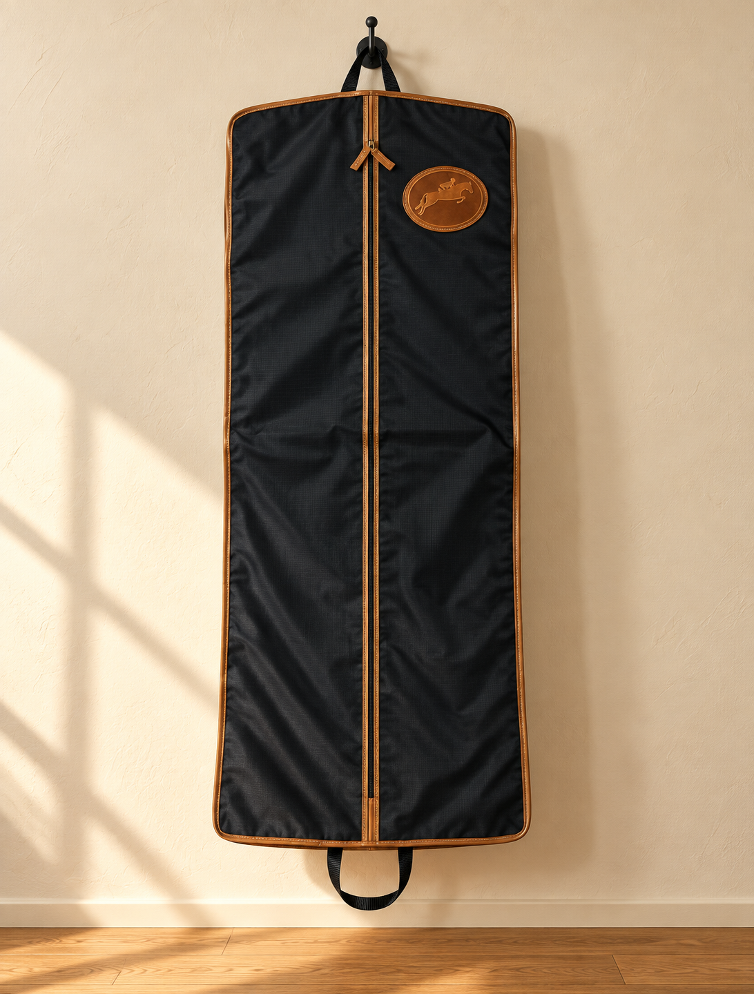 Tryon garment bag hanging from an iron hook on a cream plaster wall, with tan leather binding, a full-length centre zip and an oval embossed leather medallion