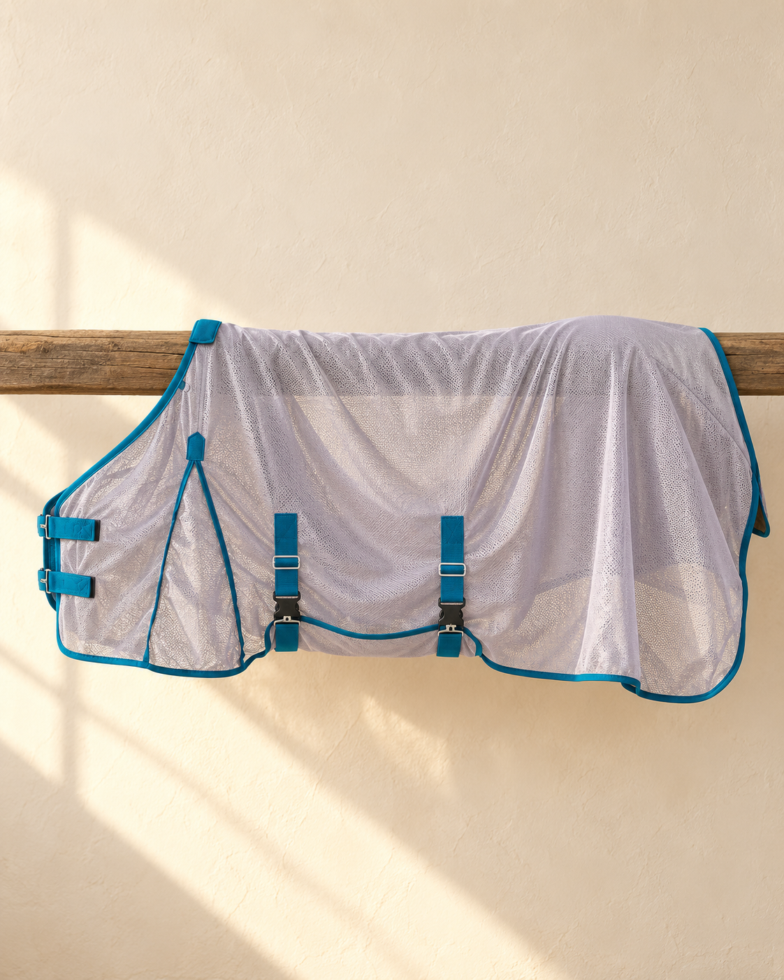 Pale grey mesh fly sheet folded over a weathered wooden rail against a cream plaster wall, showing the teal bound edges, the two surcingle straps with clip buckles and the teal chest closure
