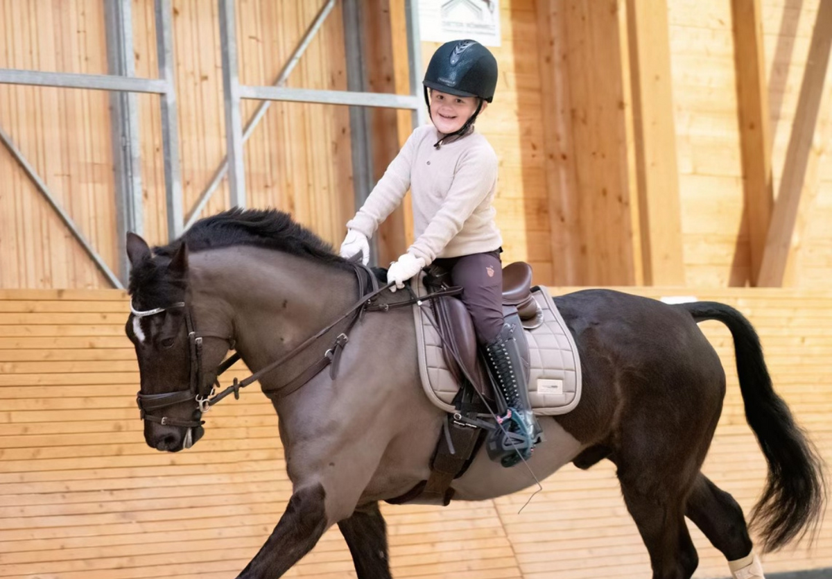 Child rider in a helmet trotting a light brown pony fitted with a neck strap in an indoor arena
