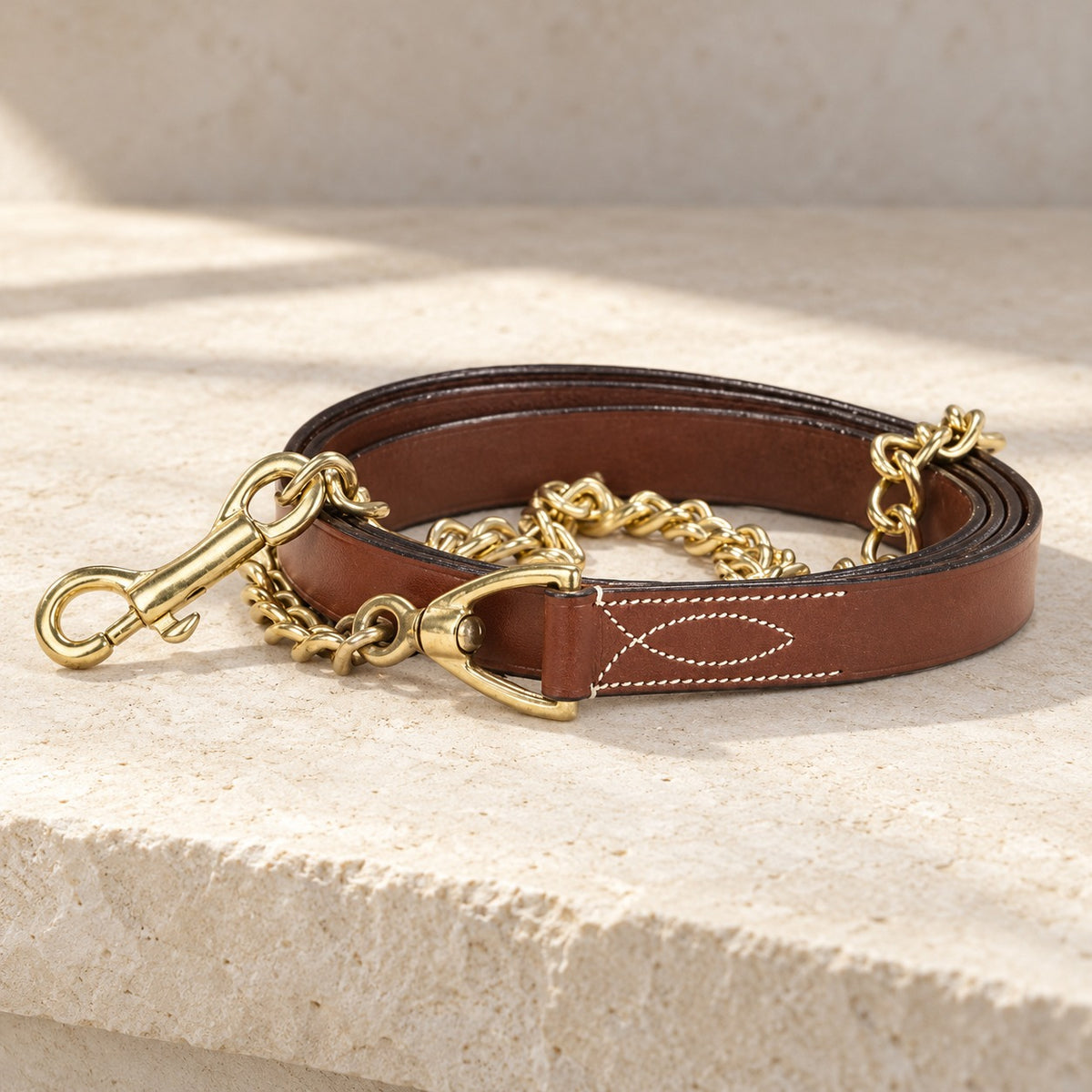 Brown leather lead with white decorative stitching and a brass chain and snap, coiled on a pale limestone ledge